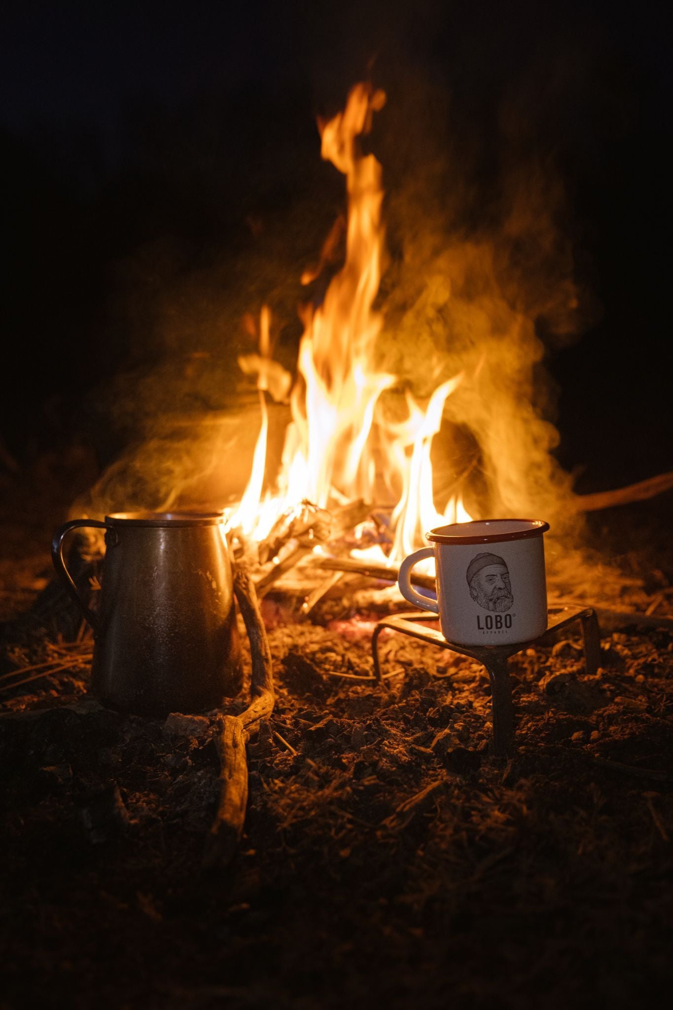 Lobo Enamel Mug featuring the bearded man with a beanie and pipe logo, warming by a glowing campfire.