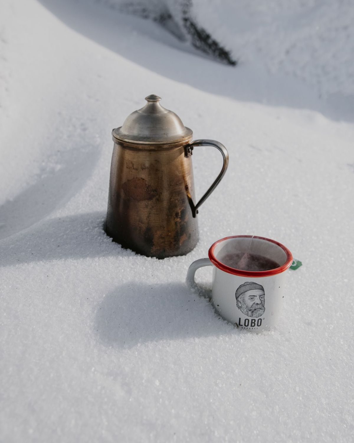 Close-up of the white Lobo Enamel Mug with hot tea in the snow, showcasing the logo of the bearded man with a beanie and pipe next to a vintage coffee pot.