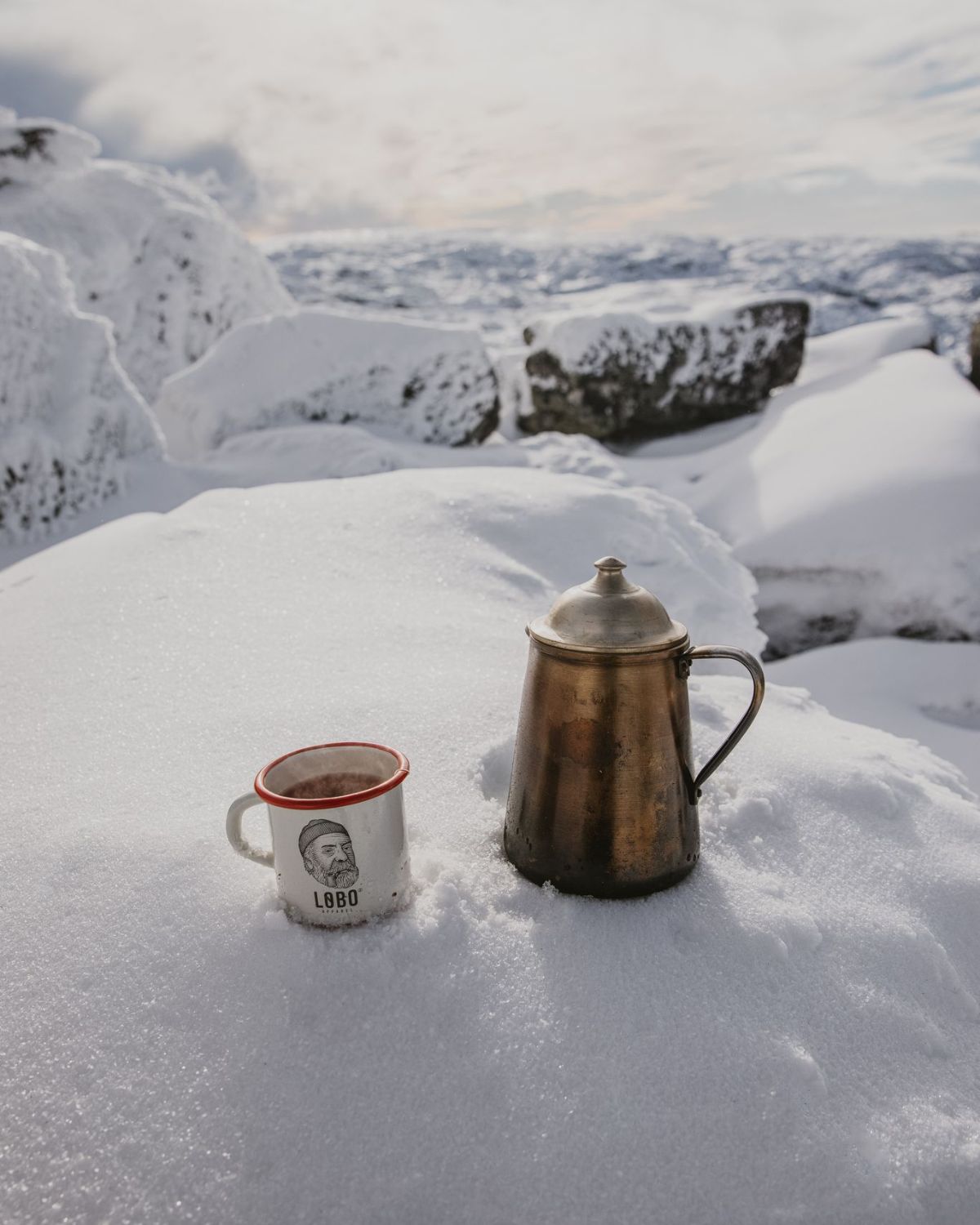 Snow-covered mountain landscape featuring the Lobo Enamel Mug and a vintage metal coffee pot, evoking outdoor adventure.