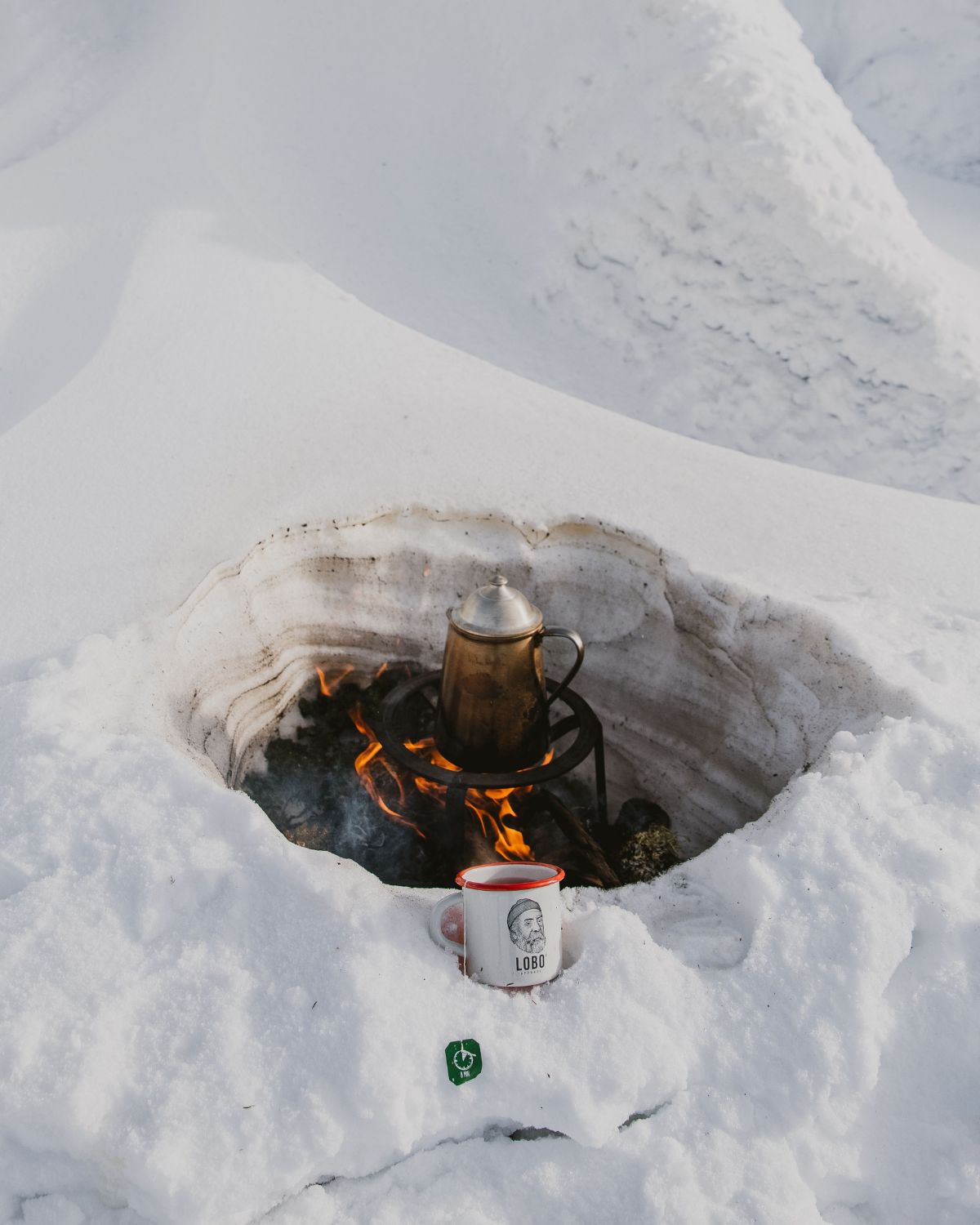 Lobo Enamel Mug resting on the snow by a campfire sheltered in an ice pit, with a metal coffee pot heating over the flames.