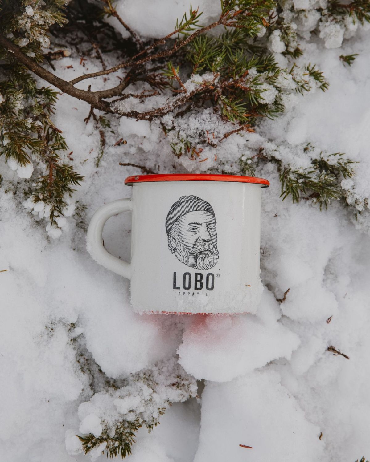 White enamel Lobo Enamel Mug highlighting the bearded man with a beanie and pipe logo, tucked into fresh snow.