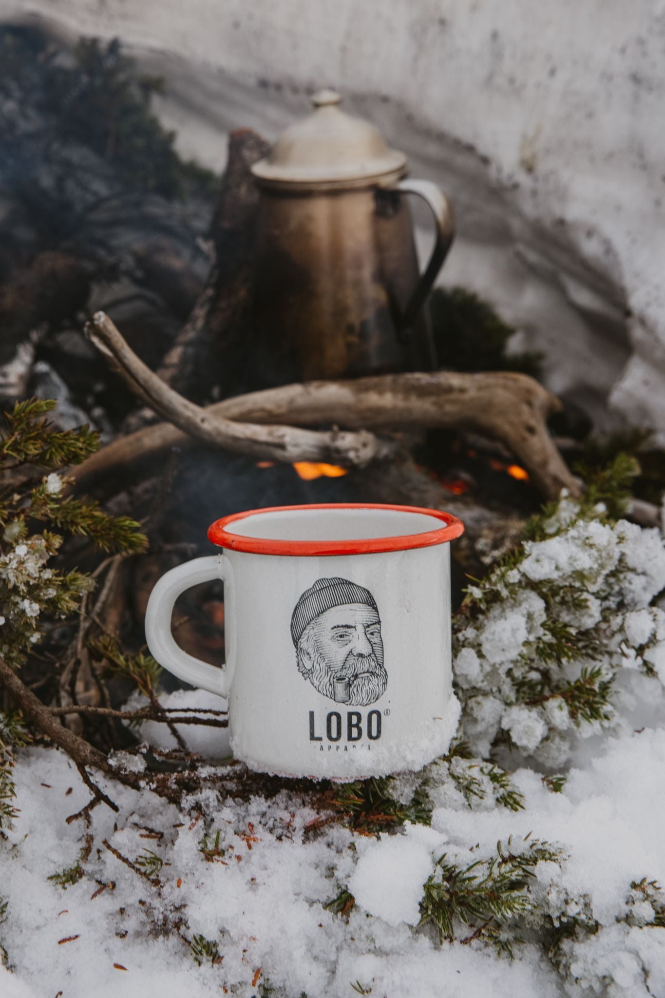 Detail of the metal Lobo Enamel Mug with a red rim, placed on snow near firewood and winter greenery.
