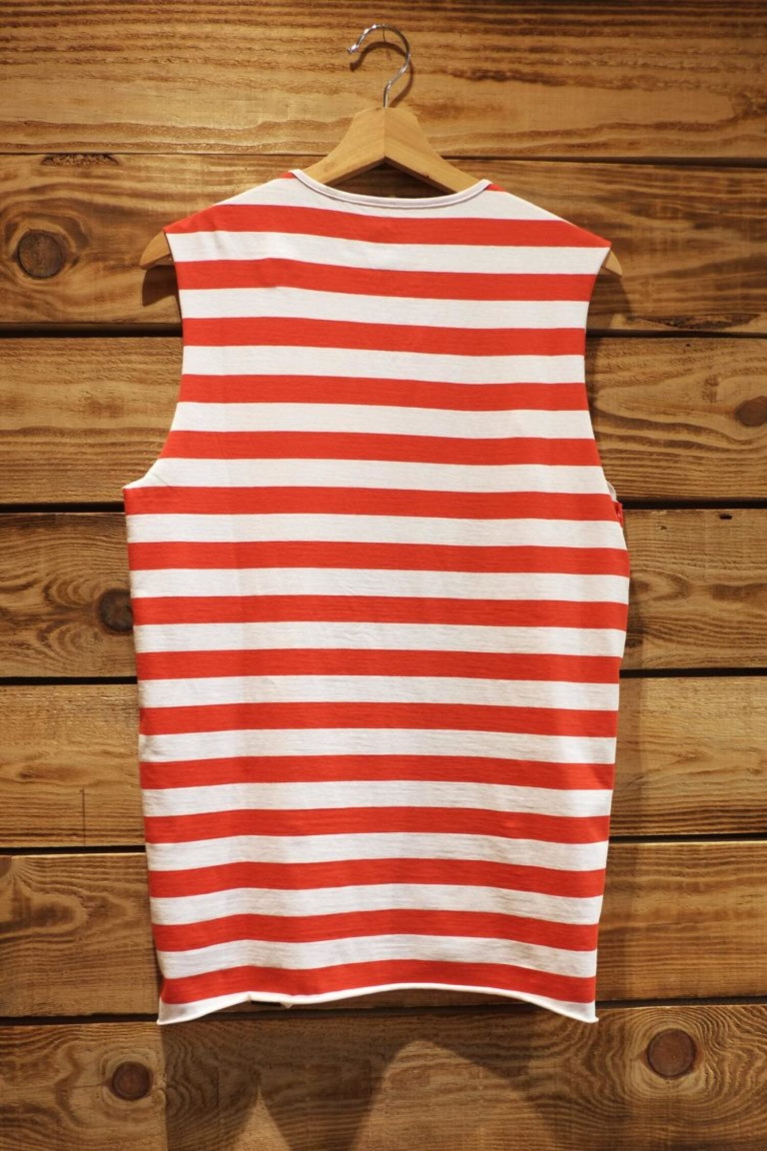 Back view of the red and white striped sleeveless t-shirt, showing the raw-edge hem finish against a rustic wooden wall background.