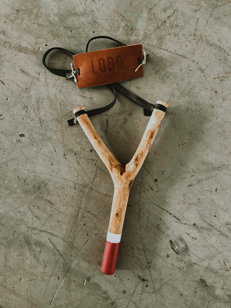 Set of exclusive LOBO x Cuscuz Studio wooden slingshots, featuring organic design and leather tags, on an industrial concrete base. Contrast between craftsmanship and modernity.