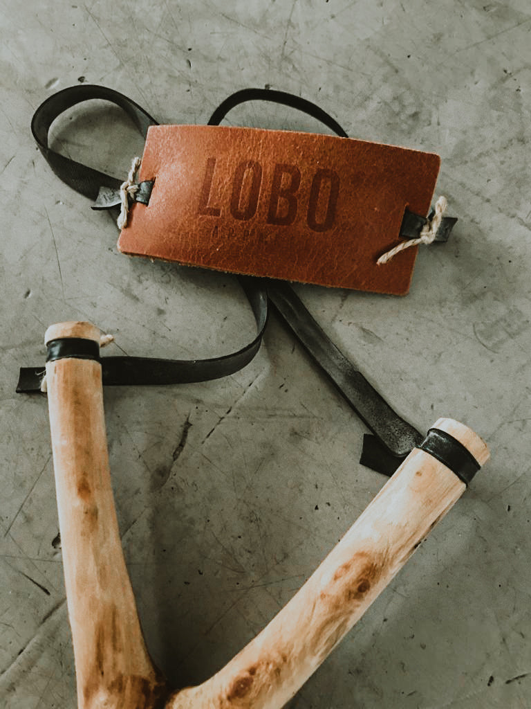 Two LOBO artisanal slingshots from the Cuscuz Studio collaboration, crafted from natural tree branches with white and red finishes, displayed on a rustic wooden counter.