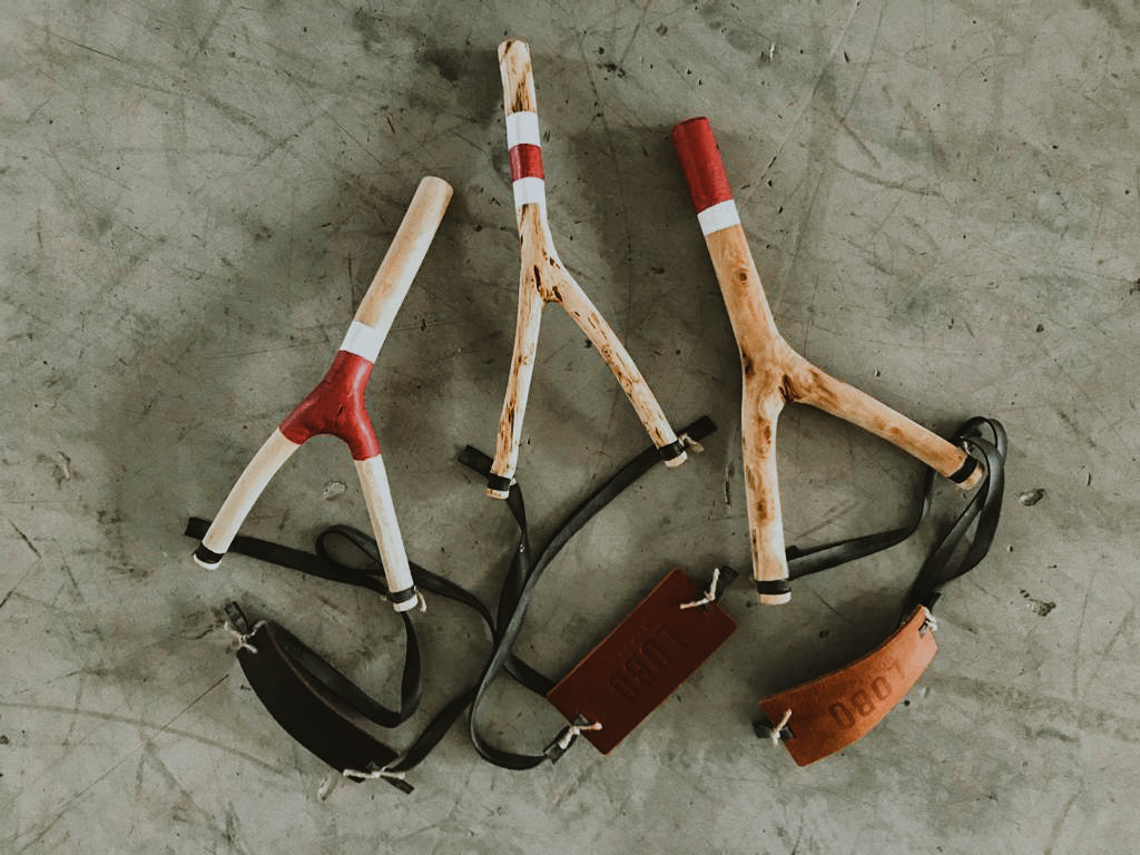 Trio of LOBO artisanal slingshots displayed on concrete and wood surfaces, highlighting the different natural branch shapes and red and white details. Features embossed leather brand tags.