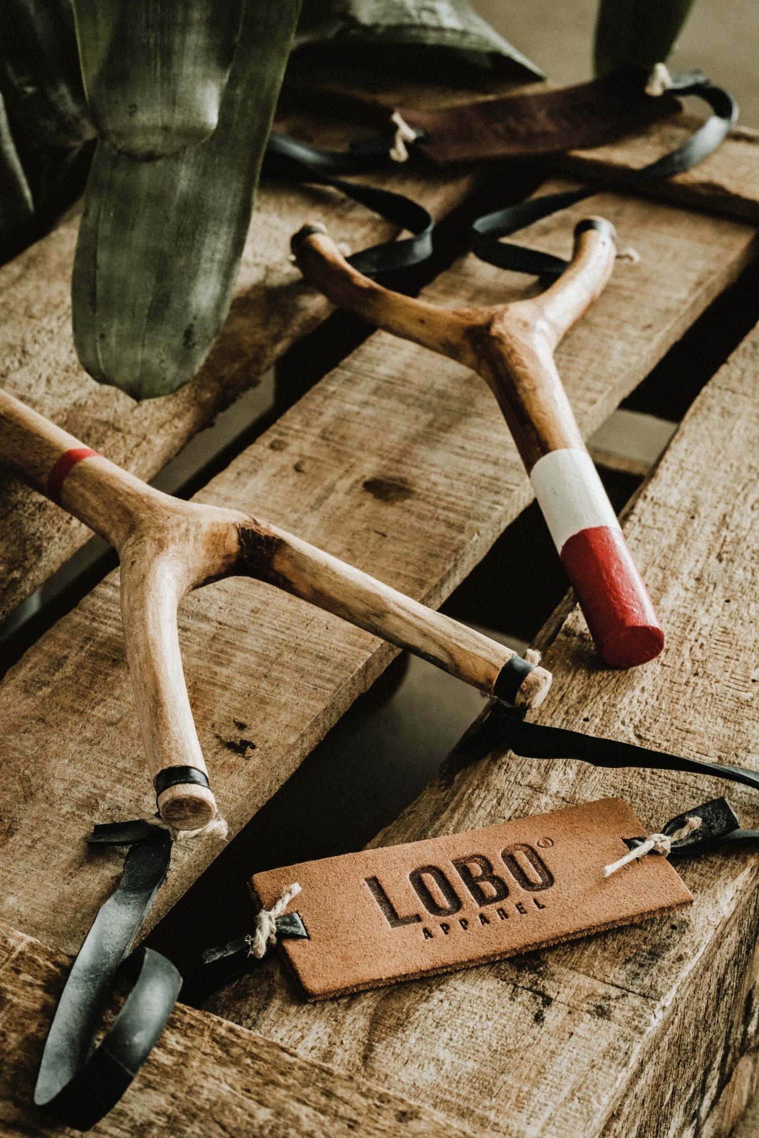 Set of LOBO artisanal slingshots, created in collaboration with Cuscuz Studio and artisan Amâncio. Natural wood handles with red and white painted details, displayed on rustic wooden pallets.