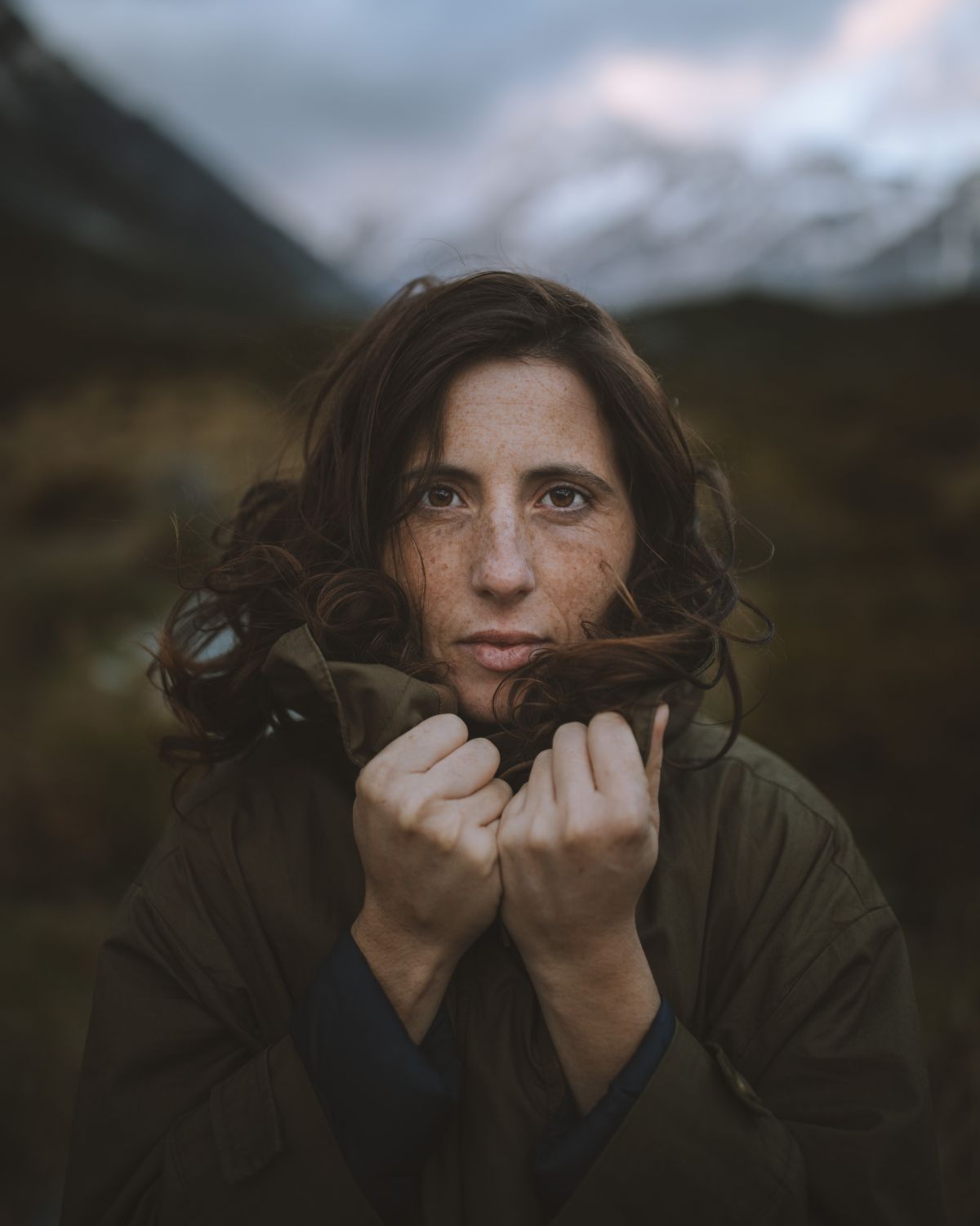 Close-up portrait of a woman with freckles and brown hair, wearing an olive green outdoor jacket in a mountain setting. Adventure lifestyle photography for LOBO brand.