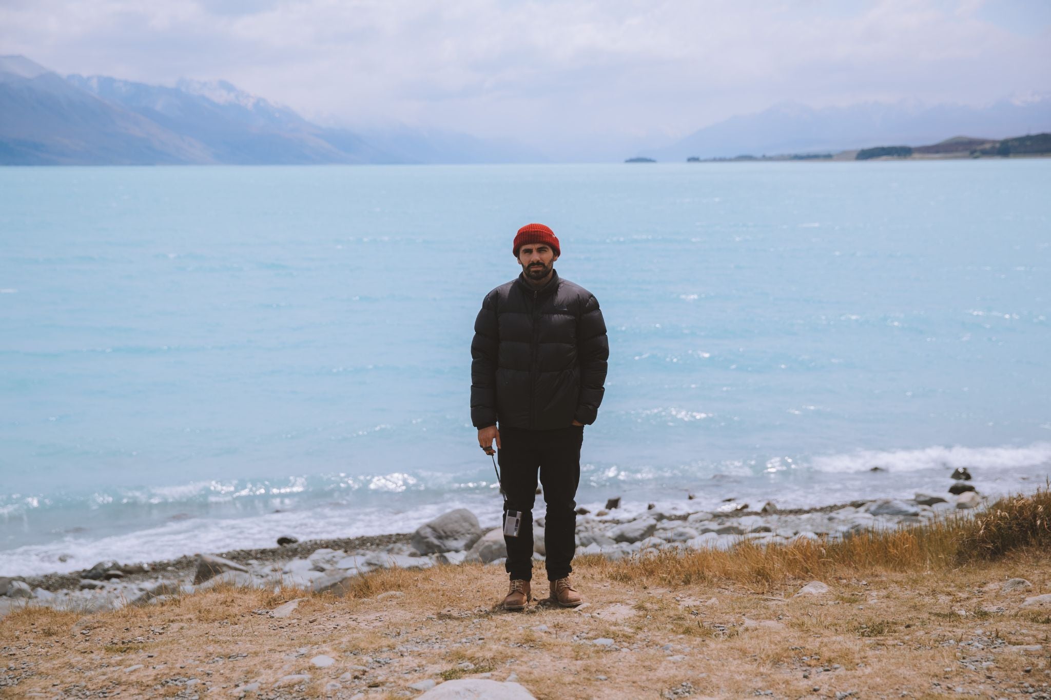 Man in Lobo Raw Boots and red beanie by a turquoise lake. Antípode collection by Tiago Sales.