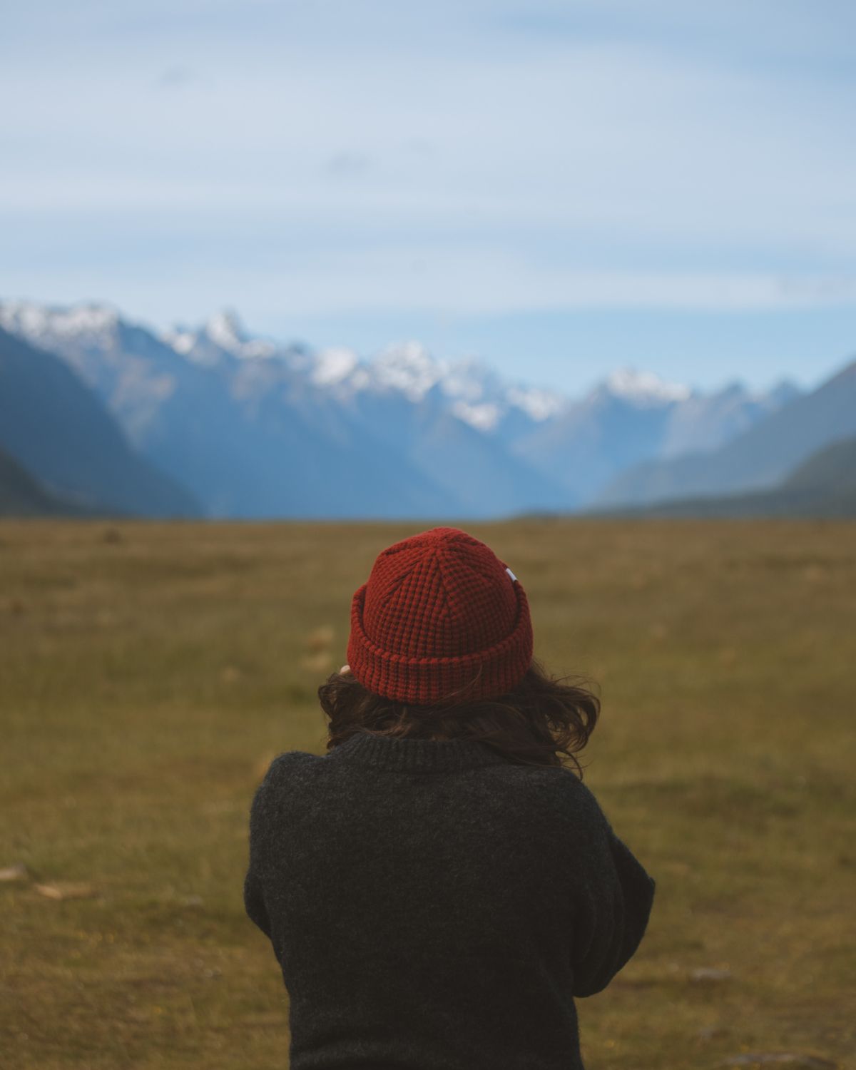 Back view of a person wearing a red beanie looking at snow-capped mountains under a cloudy sky. Nature adventure with LOBO brand.
