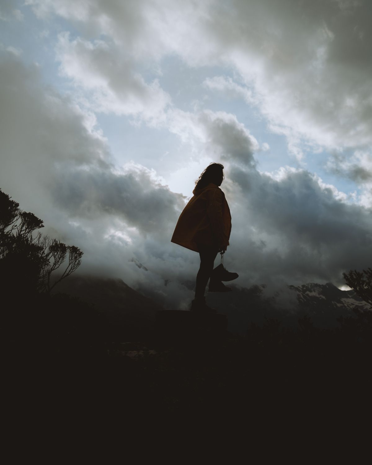 Silhouette of a person standing on rocks on a mountainside, holding a pair of Lobo Raw Boots and looking up at dramatic clouds