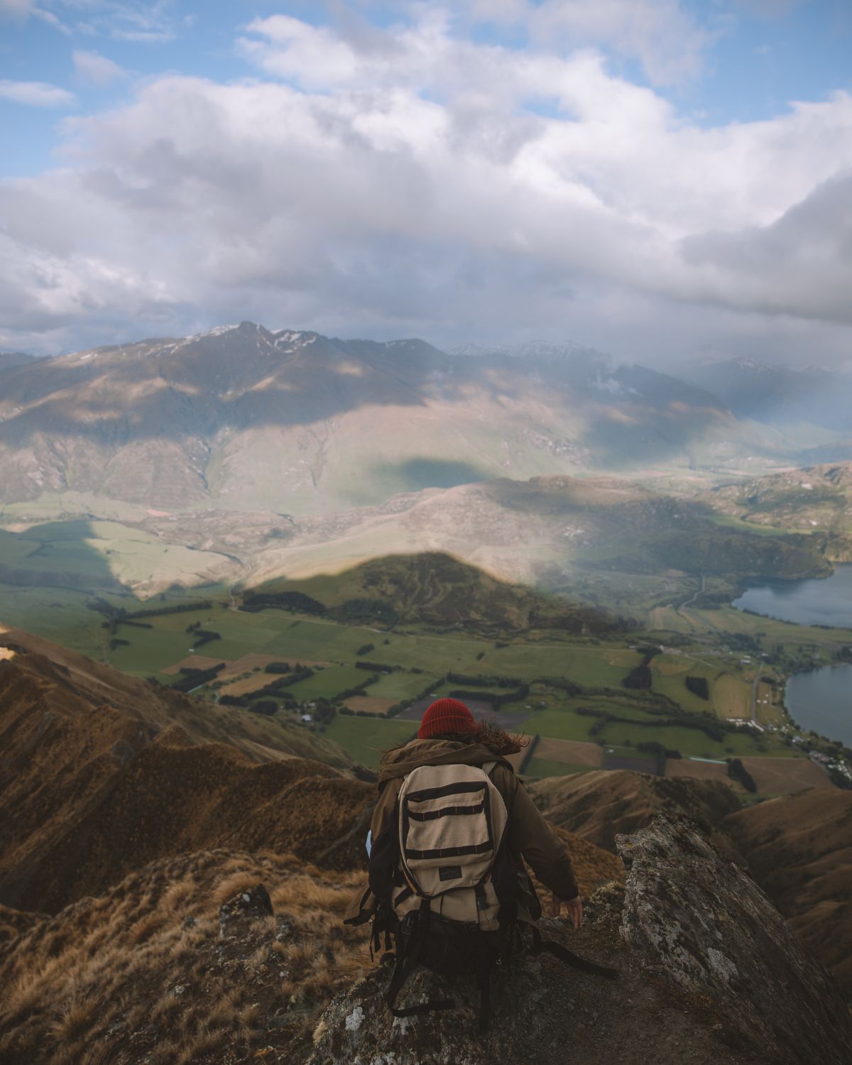 Panoramic view of a hiker with a backpack standing on a mountain peak, overlooking a green valley and distant lakes under a cloudy sky.