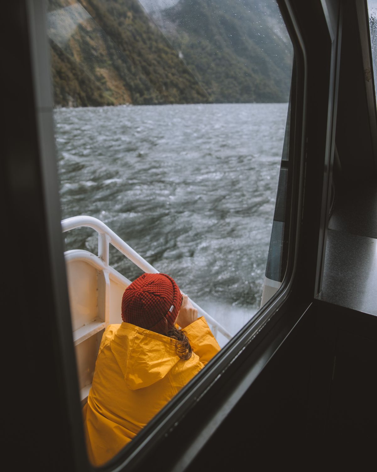 Person wearing a yellow raincoat and red beanie on a boat, overlooking lake waters and mountains. Outdoor lifestyle and adventure.