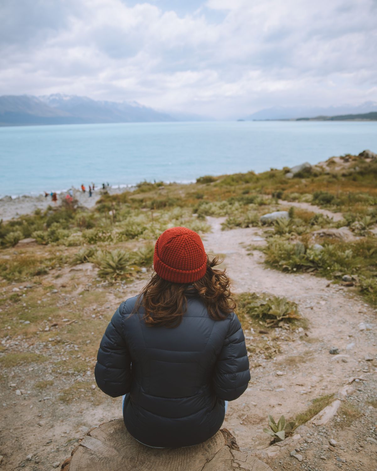 Back view of a woman in a red beanie and dark jacket looking out at a turquoise lake and mountains.