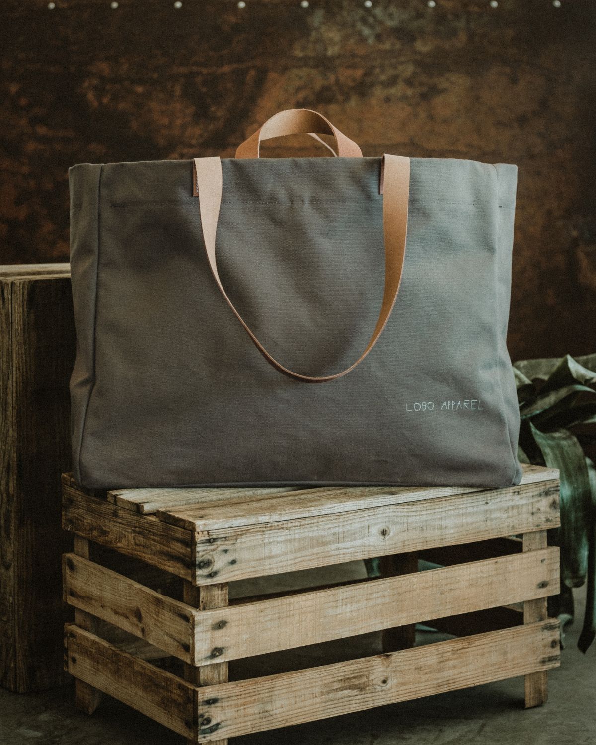 Lobo Apparel XXL military green canvas tote bag placed on a rustic wooden crate, showing leather handles and embroidered logo.