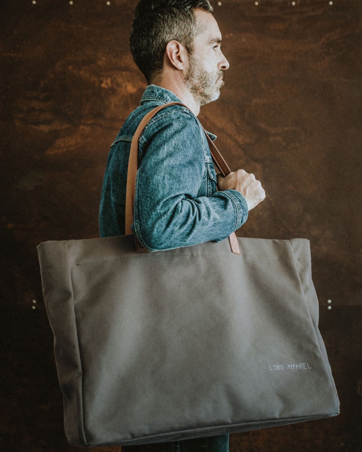 Man wearing a denim jacket carrying the Lobo Apparel XXL military green canvas bag on his shoulder, showcasing its scale and durability.