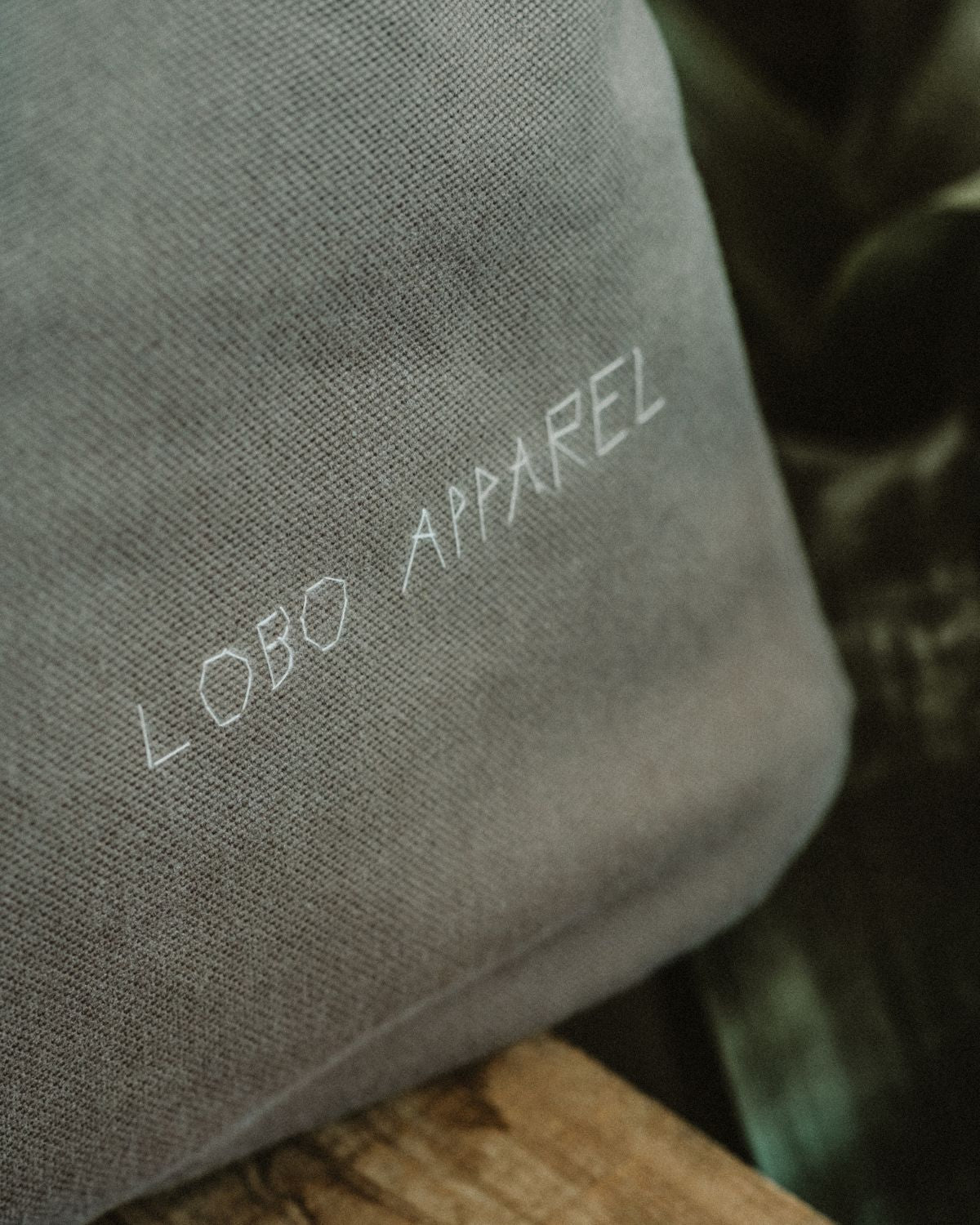 Close-up of the LOBO APPAREL logo hand-embroidered in white thread on greyish-green cotton canvas, highlighting the rustic and artisanal craftsmanship.