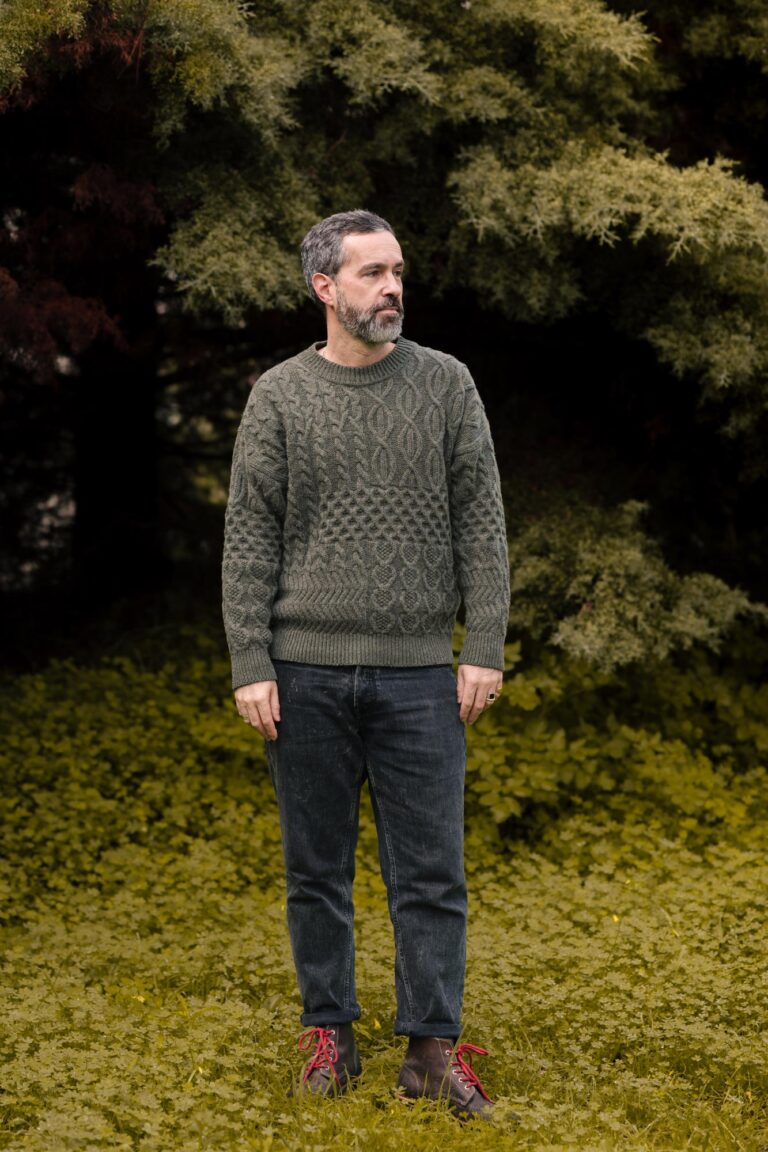 A man standing in a forest setting, wearing the "Verde Grisalho" Lobo x Do Zero unisex recycled knit sweater and Lobo Raw Boots with red laces.