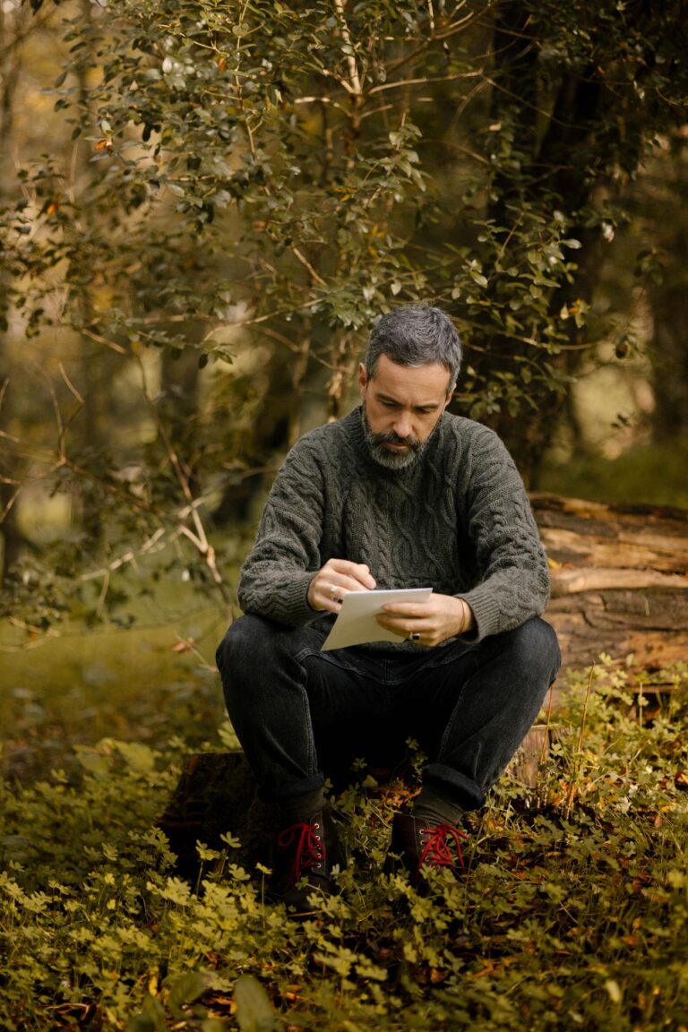 A man sitting on a log in the forest writing, wearing the Verde Grisalho Lobo x Do Zero sweater and Lobo Raw Boots leather boots with red laces.