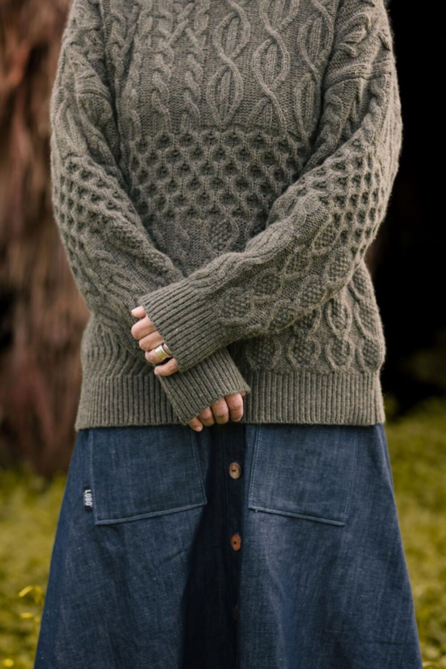 Close-up of the Verde Grisalho sweater, highlighting the intricate embossed and cable-knit patterns, paired with a long denim skirt.