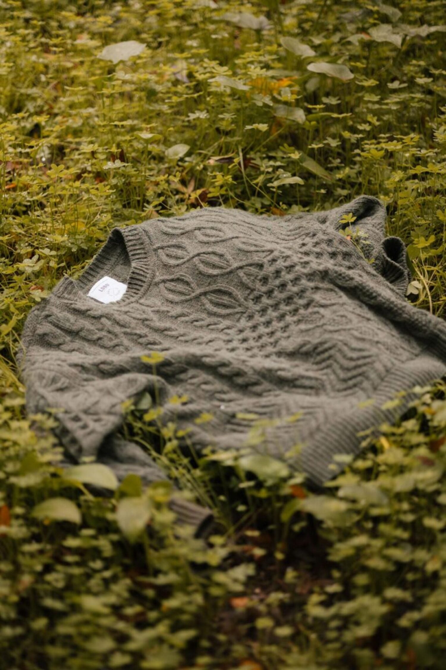 Verde Grisalho knit sweater by Lobo Apparel laid out on forest ground vegetation, highlighting its intricate cable-knit design.
