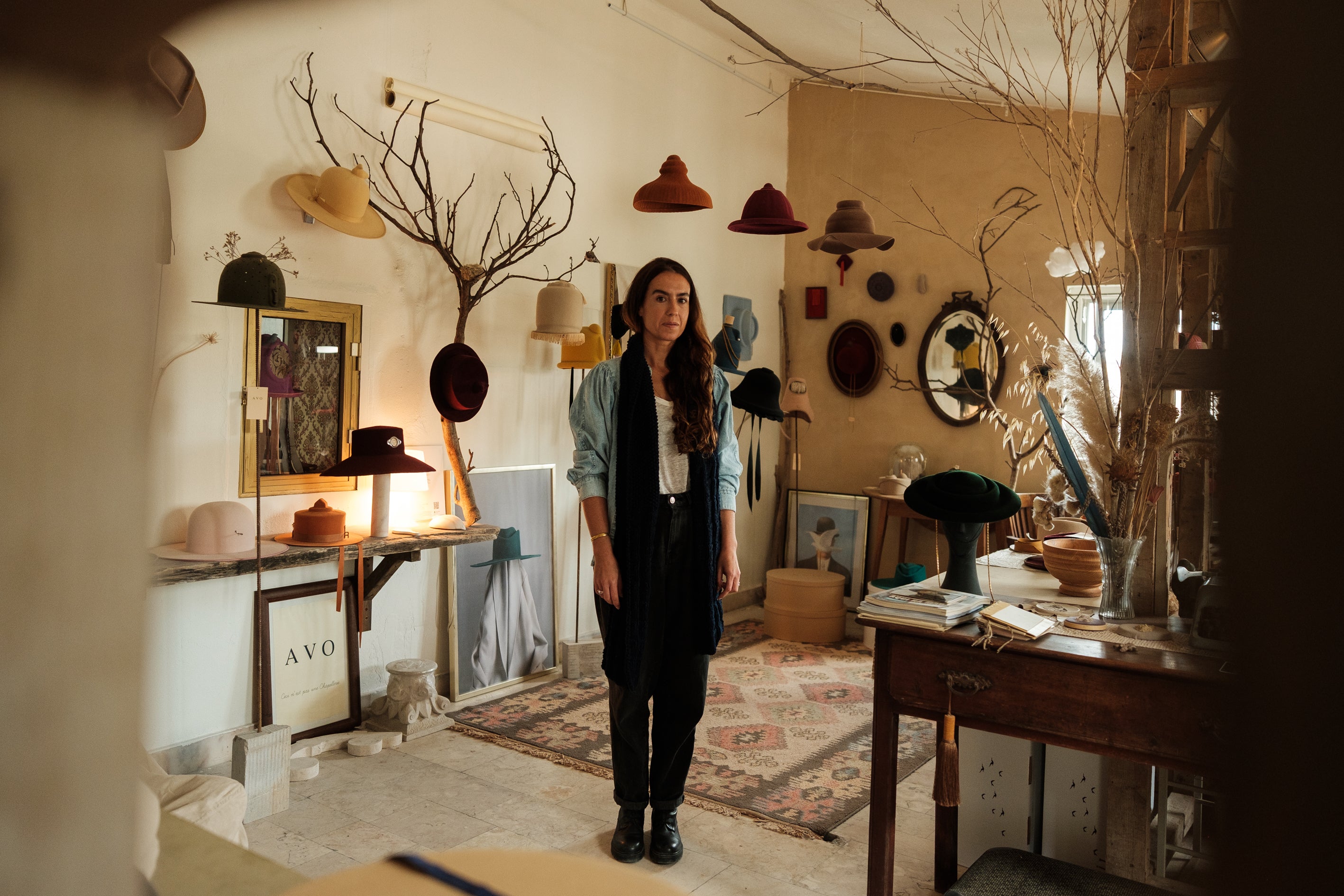 Milliner Sofia Avo standing in her Lisbon studio surrounded by handcrafted felt hats displayed on tree branches and wooden shelves. A rustic, artistic workspace showcasing bespoke hat-making and artisanal design.