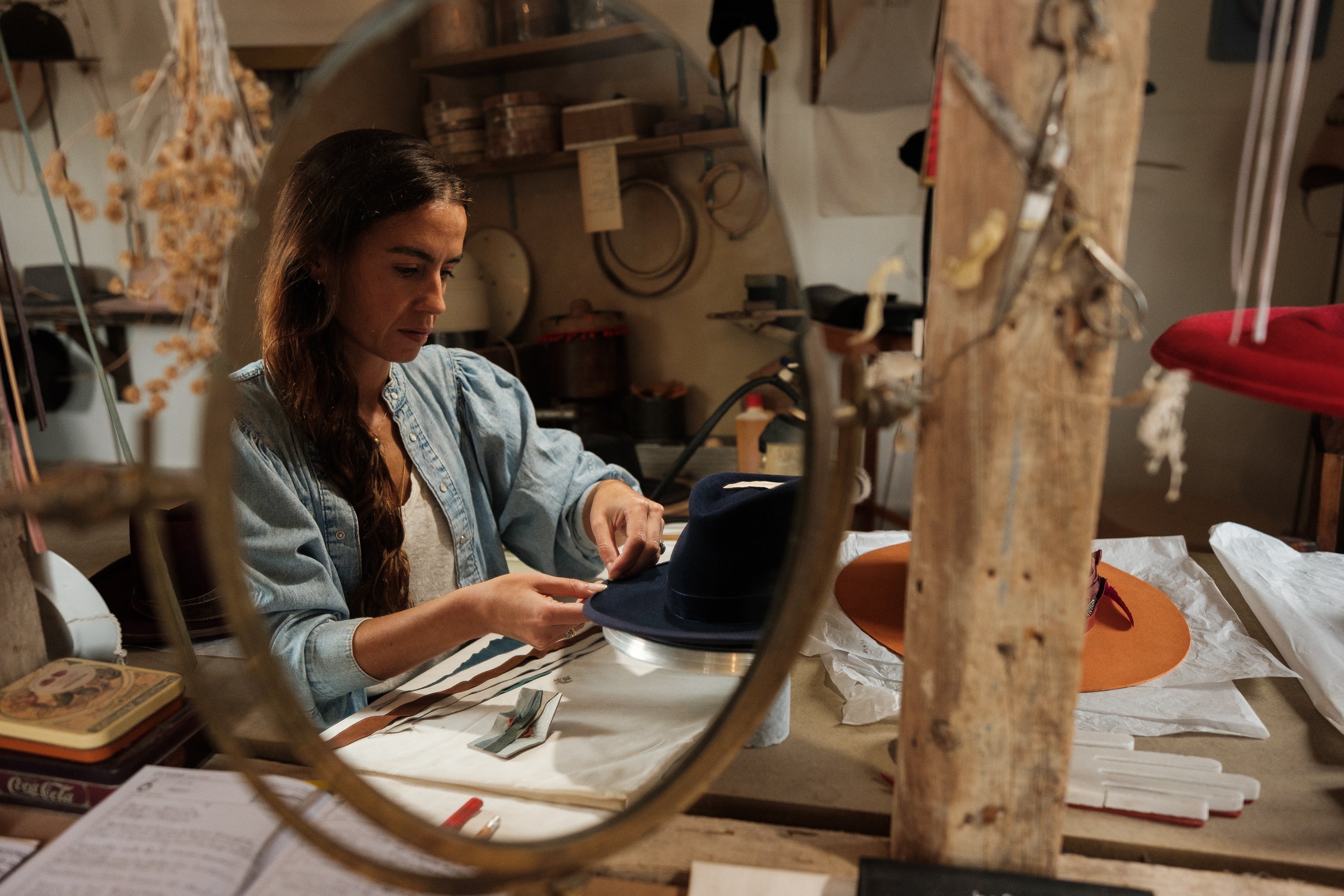 Close-up of artisan Sofia Avo in her studio, seen through an oval mirror reflection, meticulously working on a navy blue felt hat. In the background, shelves with millinery tools and hat blocks highlight the traditional craftsmanship and bespoke hat design process in Lisbon.