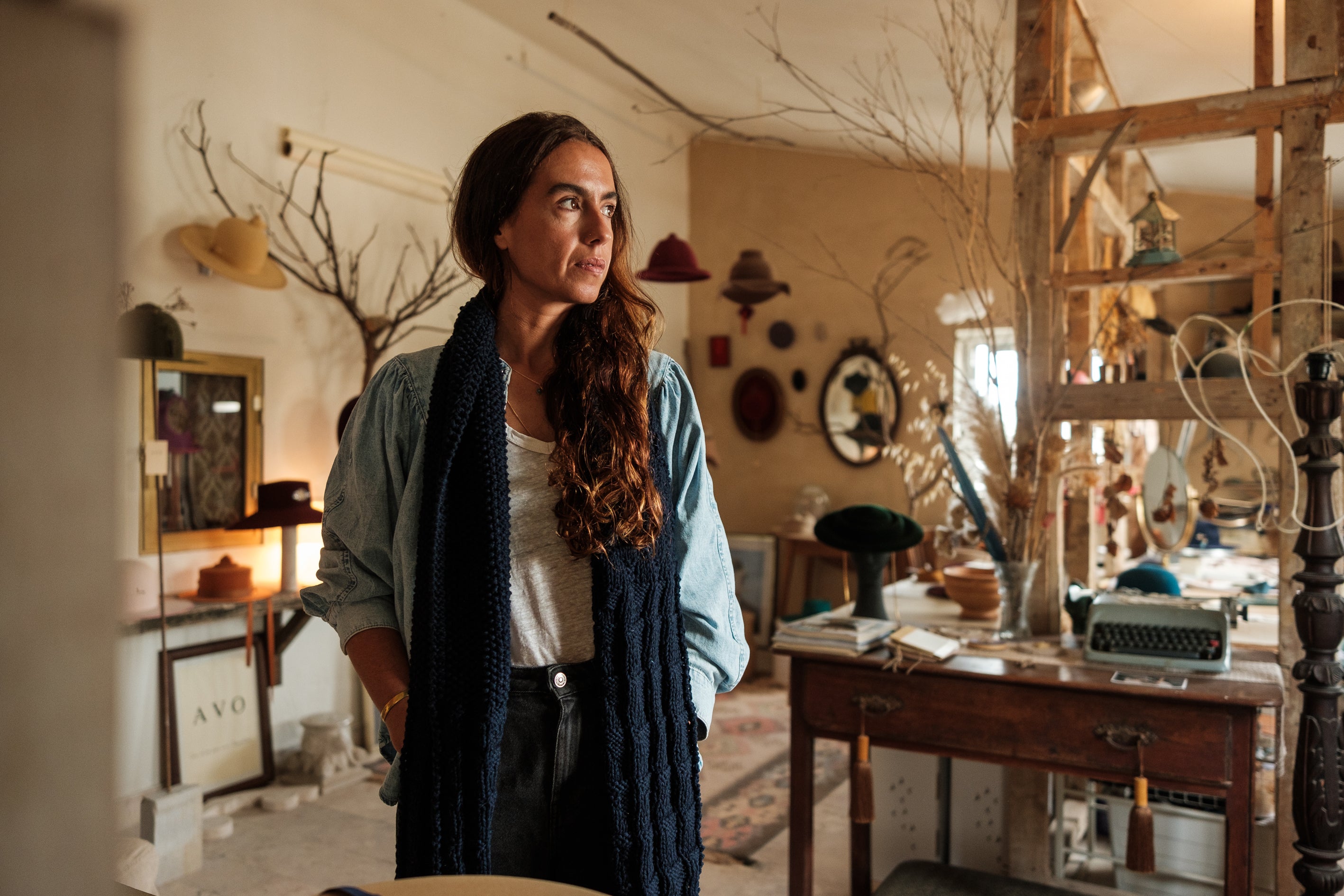 Contemplative portrait of milliner Sofia Avo in her artisanal hat studio. The scene features the creator surrounded by colorful felt hats, a vintage typewriter, and natural decor, capturing the essence of bespoke design and slow craftsmanship in Lisbon.