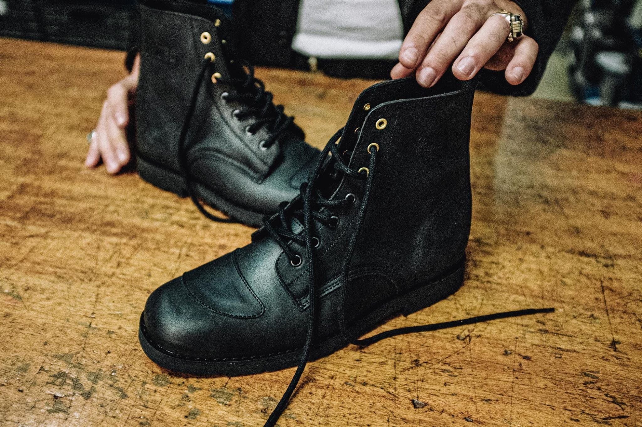 Black leather Lobo Tigre Boots on a wooden workbench, featuring an artisan's hand touching the boot collar, highlighting the golden eyelets and the embossed brand logo.