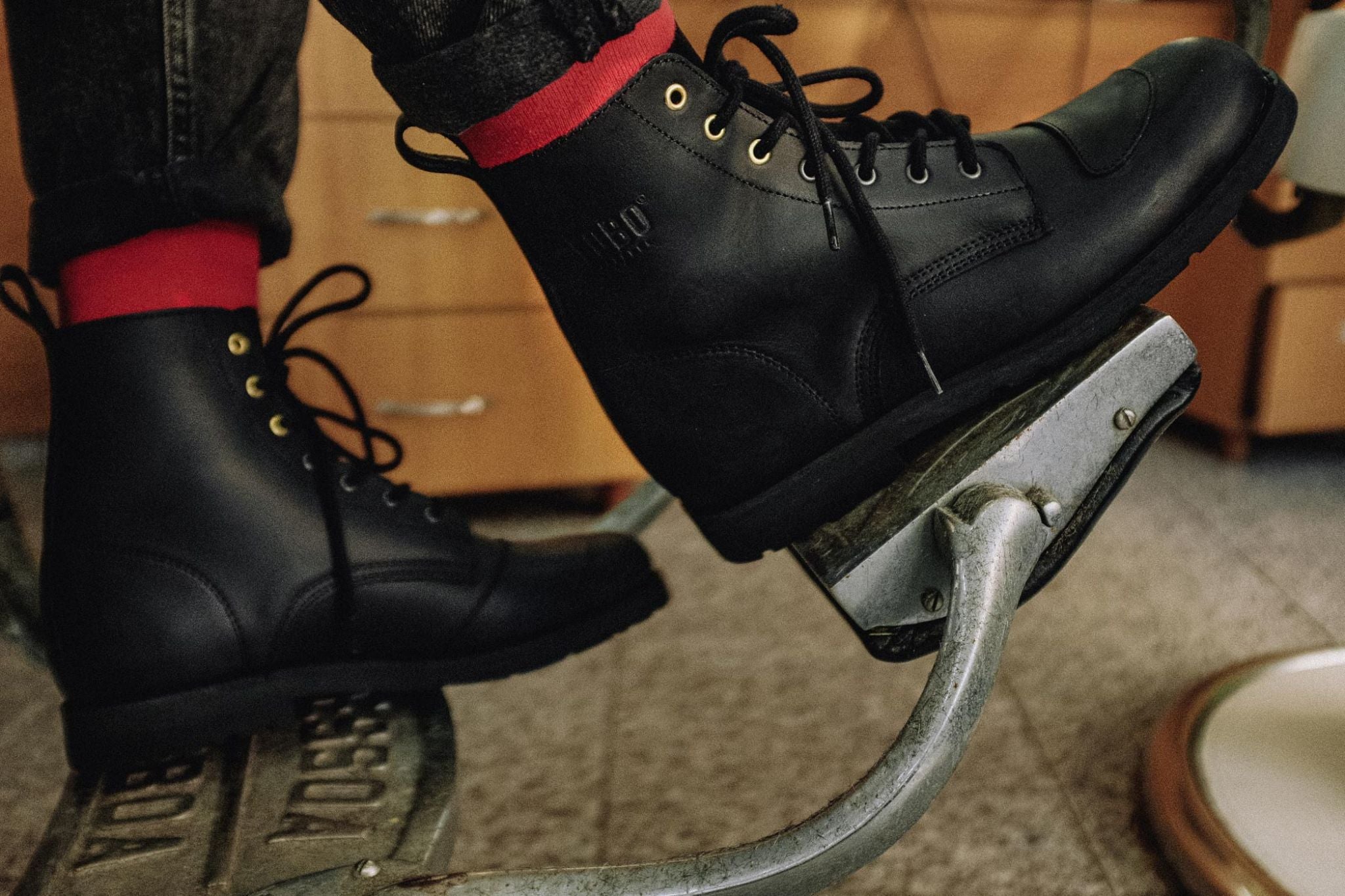Lobo Tigre Boots in black leather featuring toe reinforcement for motorcycle shifter protection and golden eyelets, resting on a metal pedal.