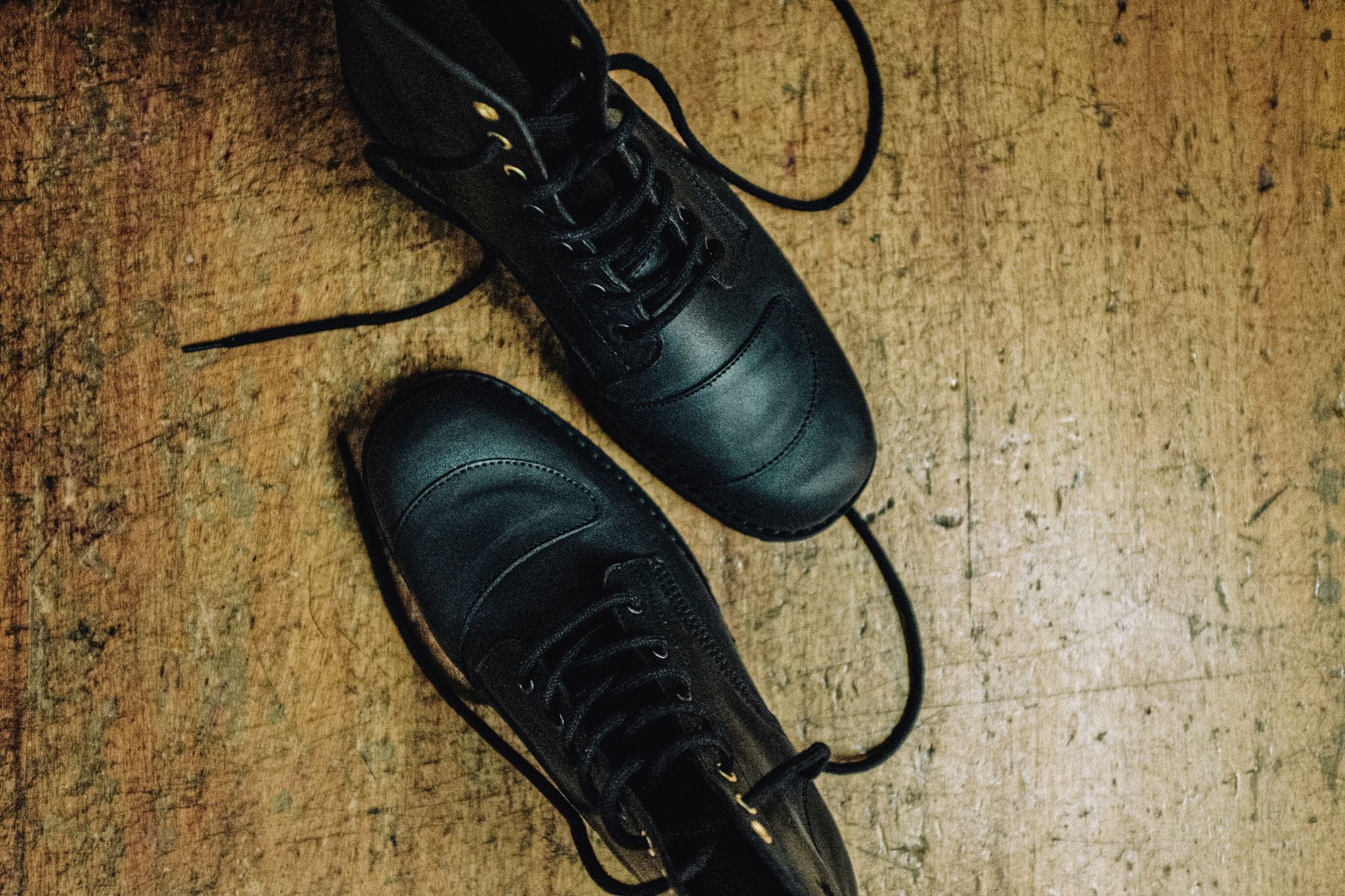 Top-down view of black leather Lobo Tigre Boots on a rustic wooden surface, showcasing the shifter protector on both toe caps and black laces.