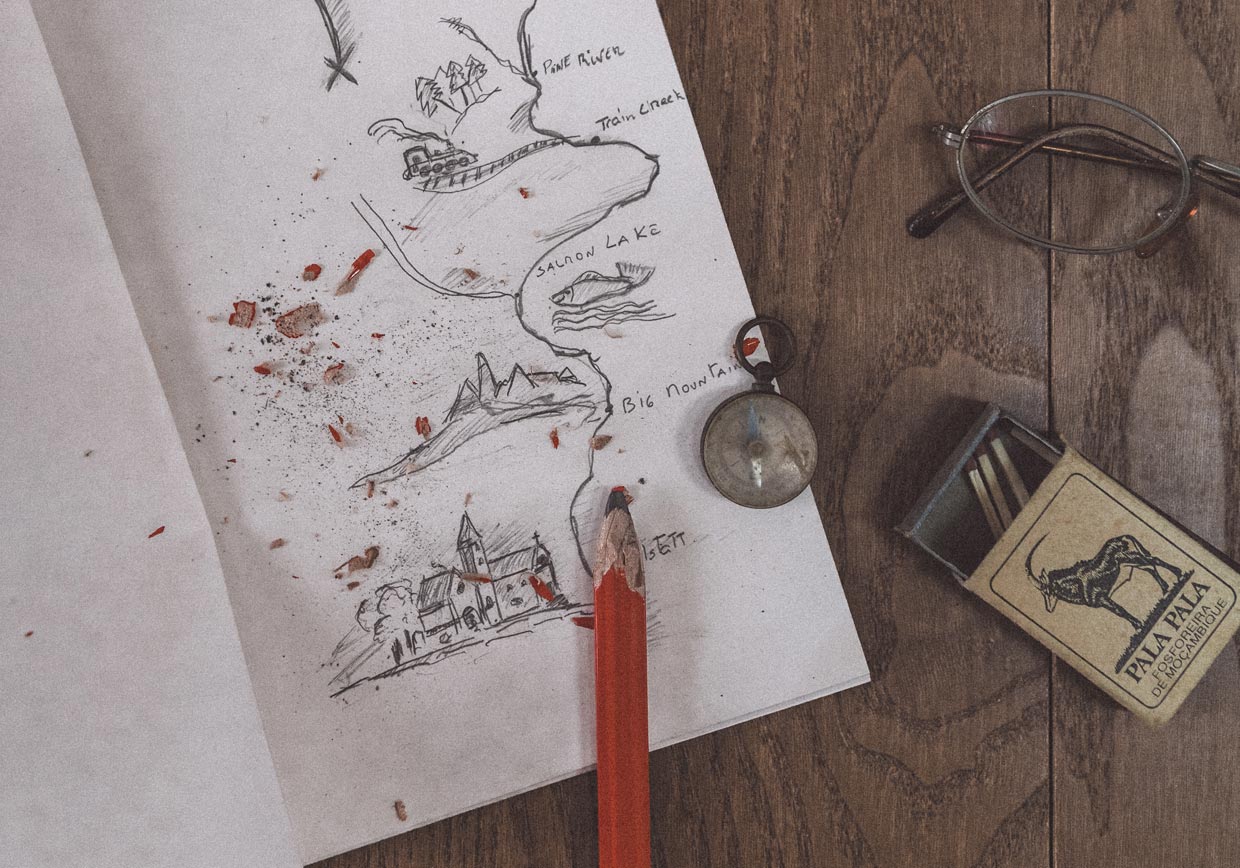 : Top view of an open notebook with a hand-drawn pencil map, illustrating travel elements like "Salmon Lake", "Big Mountain", and "Hallstatt". On a wooden table, there is a sharpened red pencil, an antique compass, a vintage Pala Pala matchbox, and reading glasses.