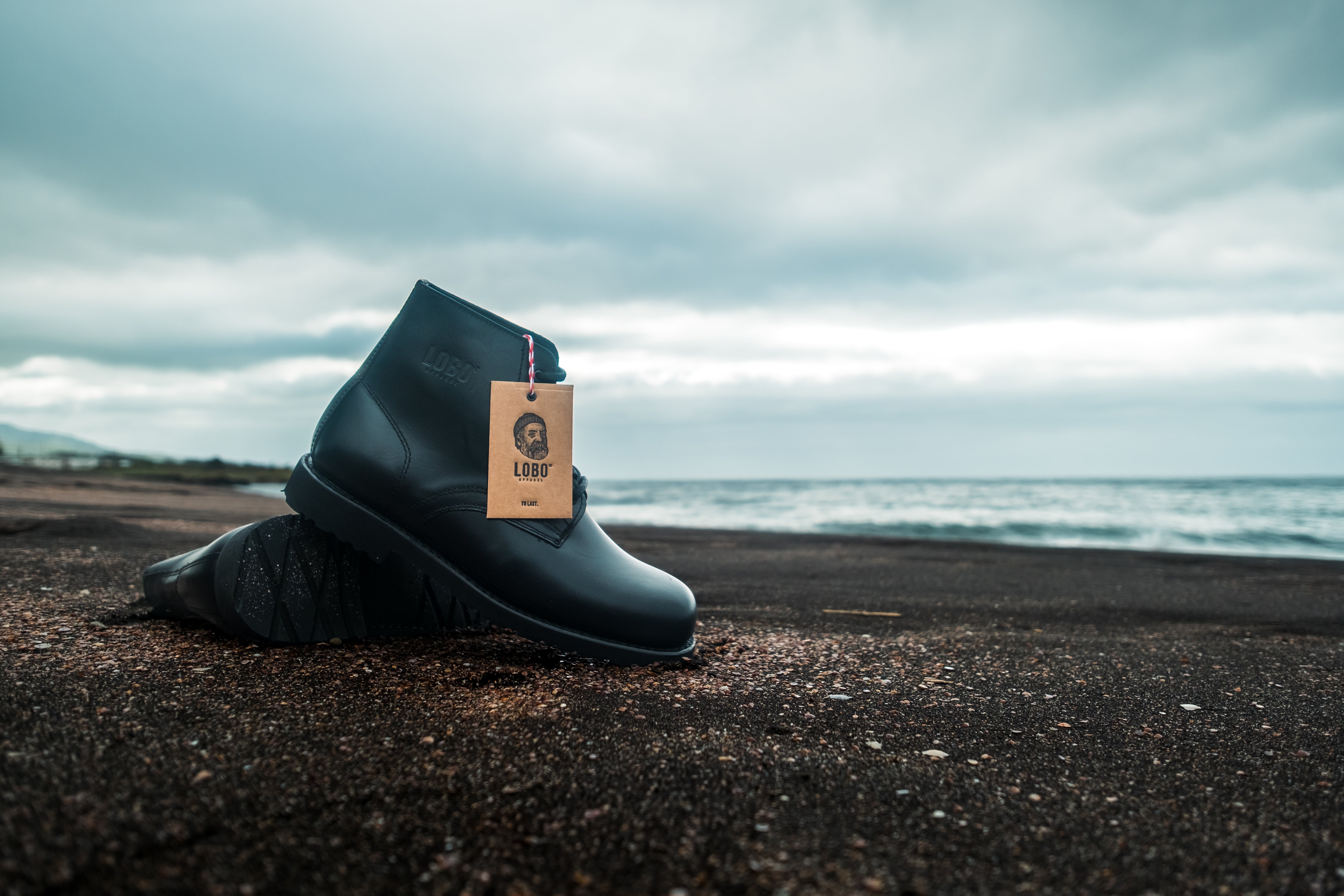 Pair of Lobo black leather boots with Goodyear welt construction, showcased on the volcanic sand of the Azores under a moody sky.