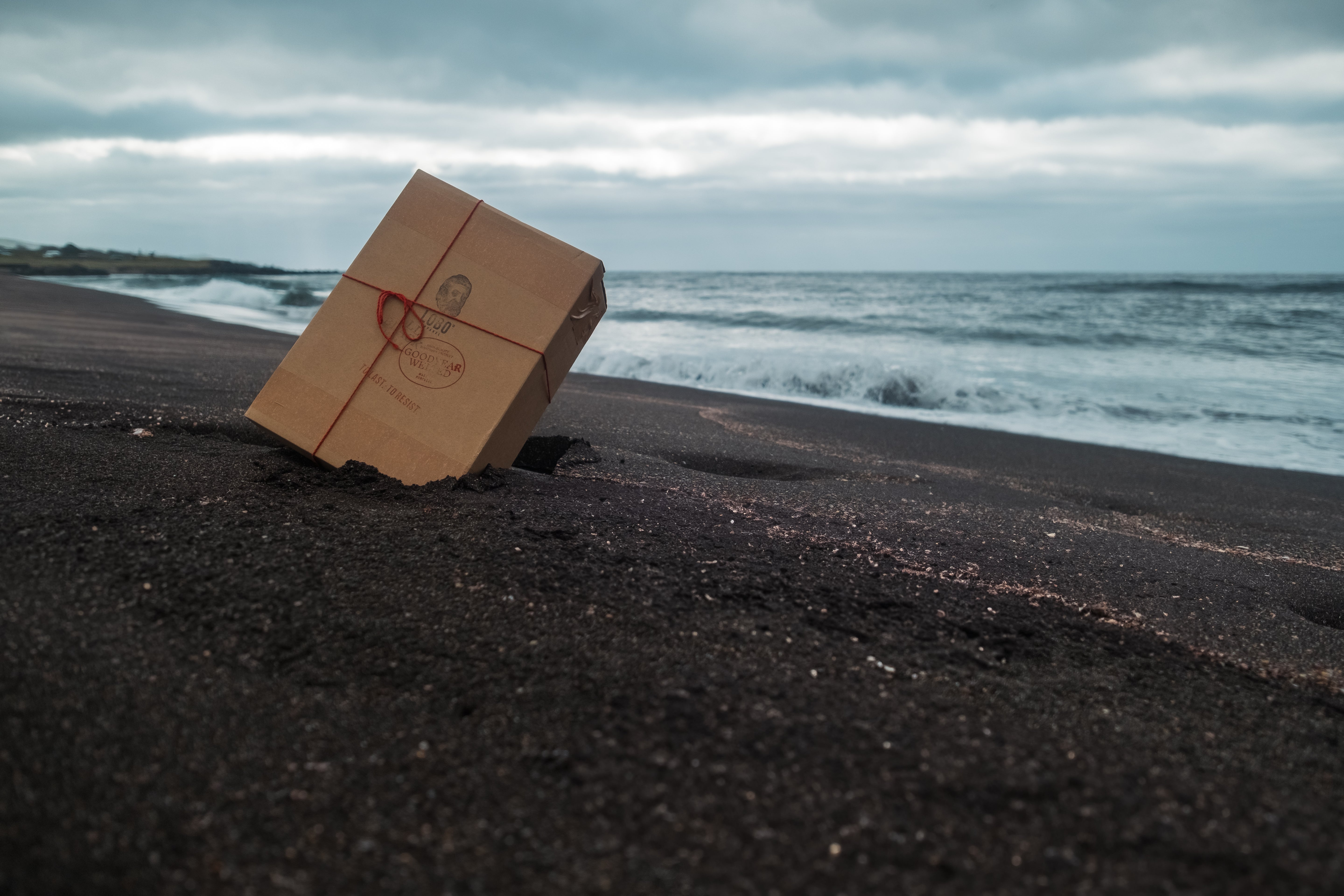 Cardboard box of All Black boots on the black volcanic sand of a beach in the Azores, with rough sea and cloudy sky in the background. Photography by Paulo Jorge.