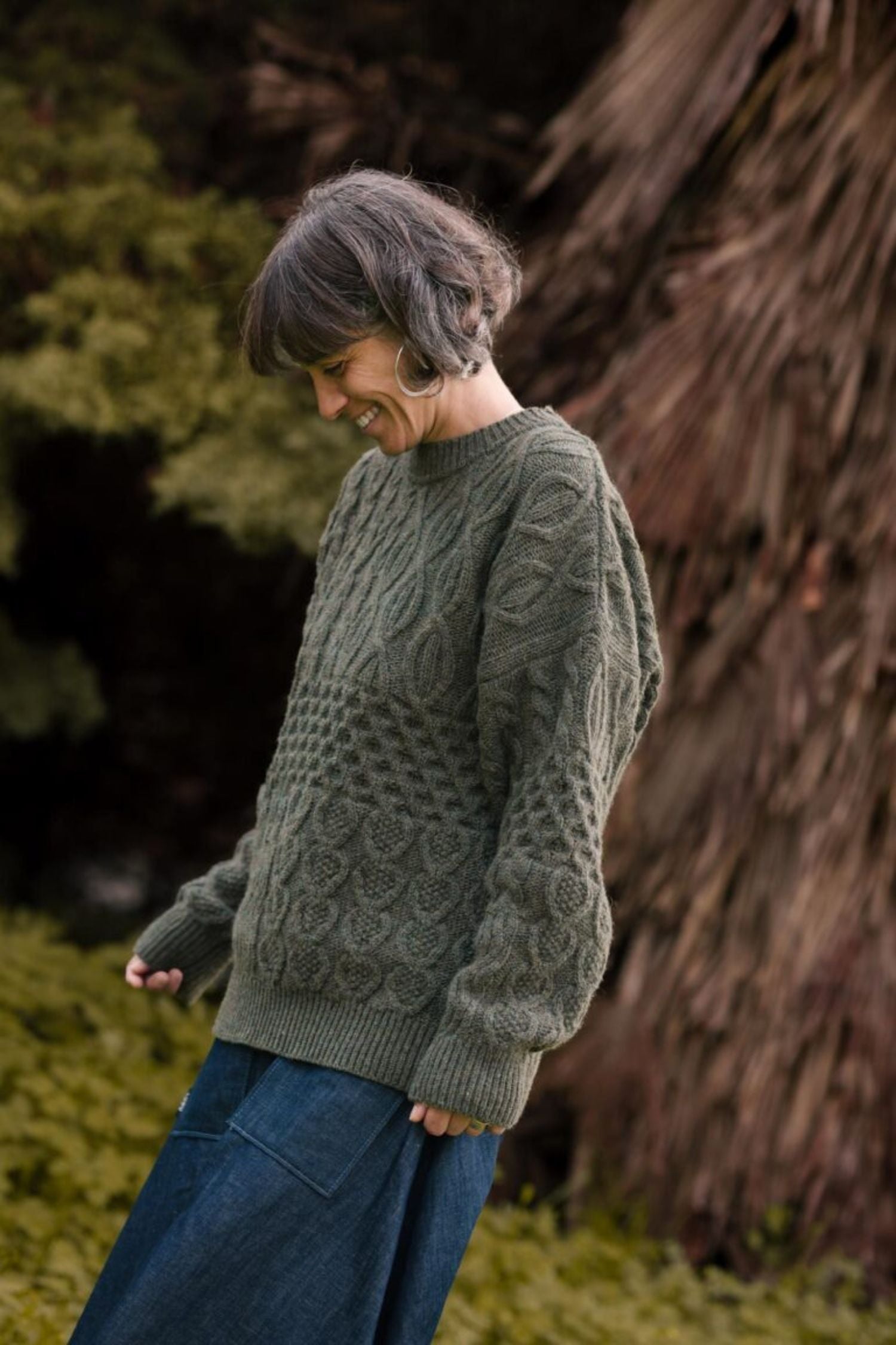 A woman smiling in profile in a natural setting, wearing the "Verde Grisalho" Lobo x Do Zero sweater made from 100% recycled knit, paired with a long denim skirt.
